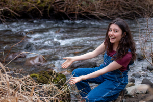 Skylar Lipman smiles while kneeling at the edge of a flowing creek with their hands stretched outwards, palms up to display a rock covered in moss.